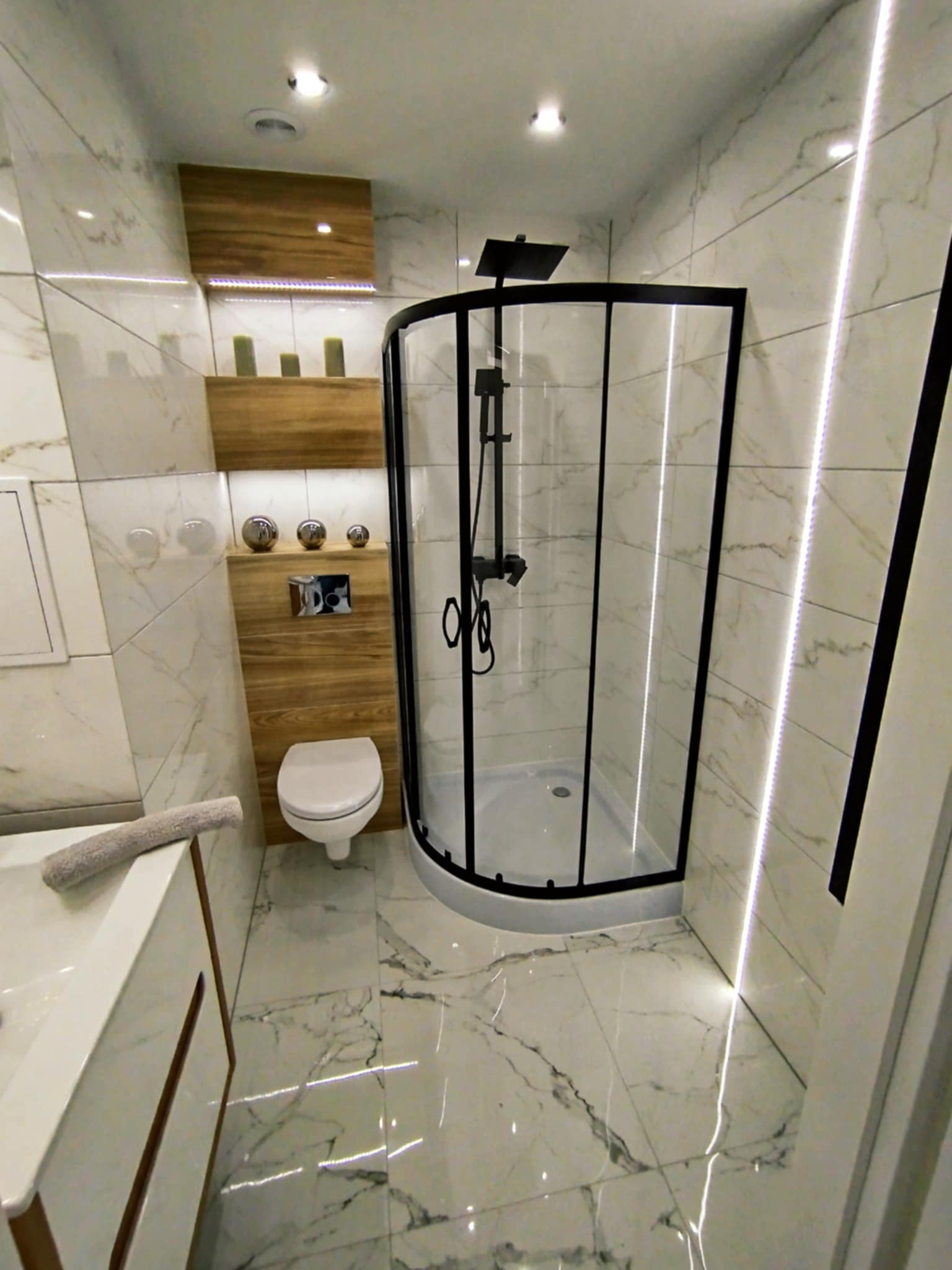Nowoczesna łazienka z kabiną prysznicową typu walk-in, marmurowymi płytkami i drewnianymi elementami dekoracyjnymi. Oświetlenie LED podkreśla elegancję wnętrza.