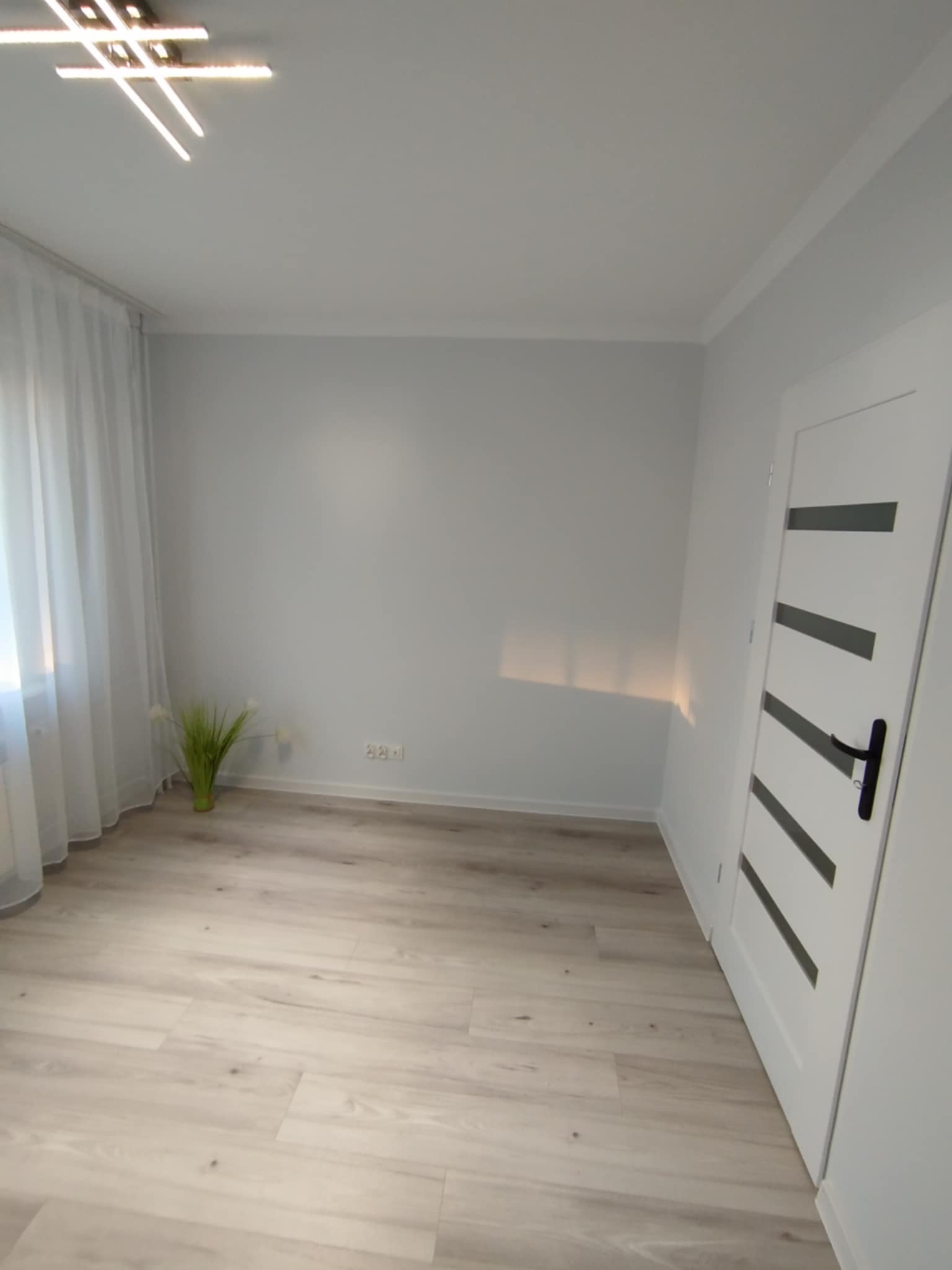 Jasne panele podłogowe w pokoju po remoncie z białą ścianą, zasłoniętym oknem i nowoczesnym oświetleniem sufitowym. Widoczne gniazdko elektryczne i białe drzwi z czarnymi akcentami.