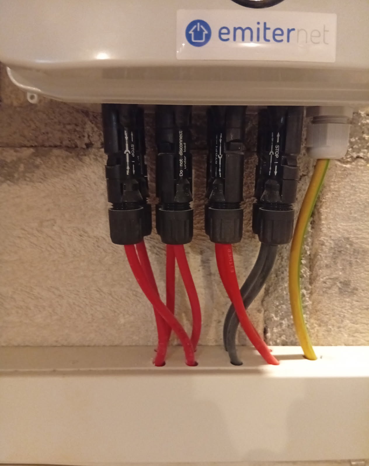 Podłączenie przewodów do instalacji fotowoltaicznej wewnątrz skrzynki. Widoczne złącza, kable czerwone, szare i żółto-zielony. Etykieta 'emiternet' na obudowie.