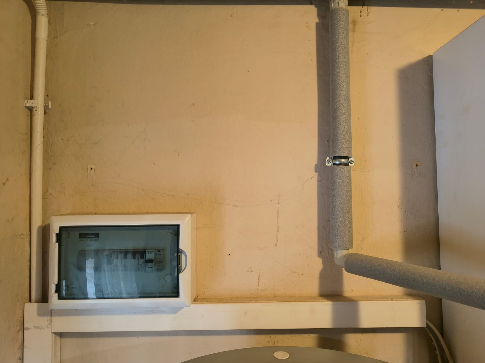 Zamknięta skrzynka elektryczna z bezpiecznikami na ścianie, obok rura w izolacji. Widoczne ślady użytkowania na ścianie. Ujęcie z bliska.