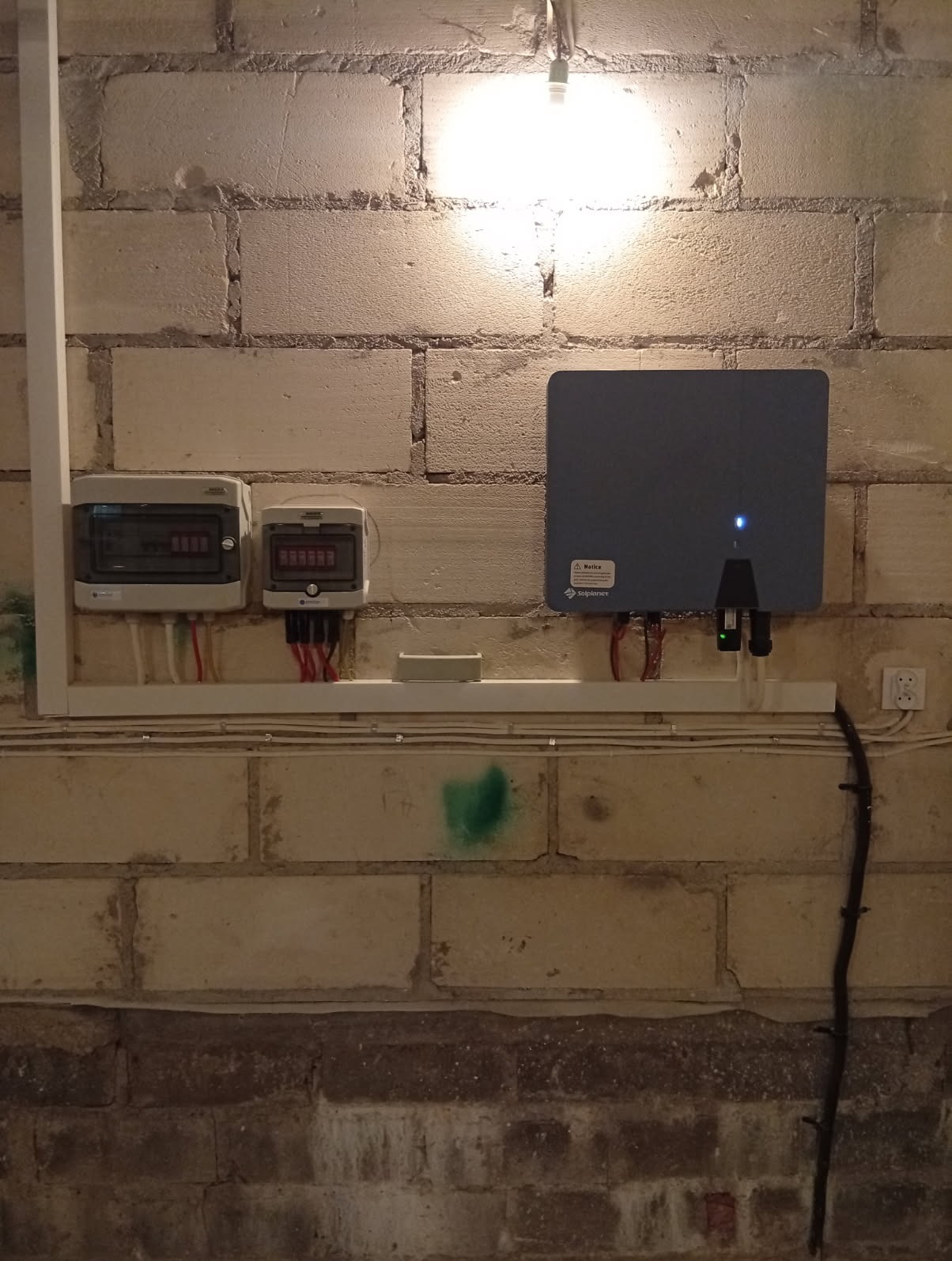 Dwa bezpieczniki i szara skrzynka elektryczna z podłączonymi kablami, zamontowane na ścianie z cegły w pomieszczeniu oświetlonym żarówką.