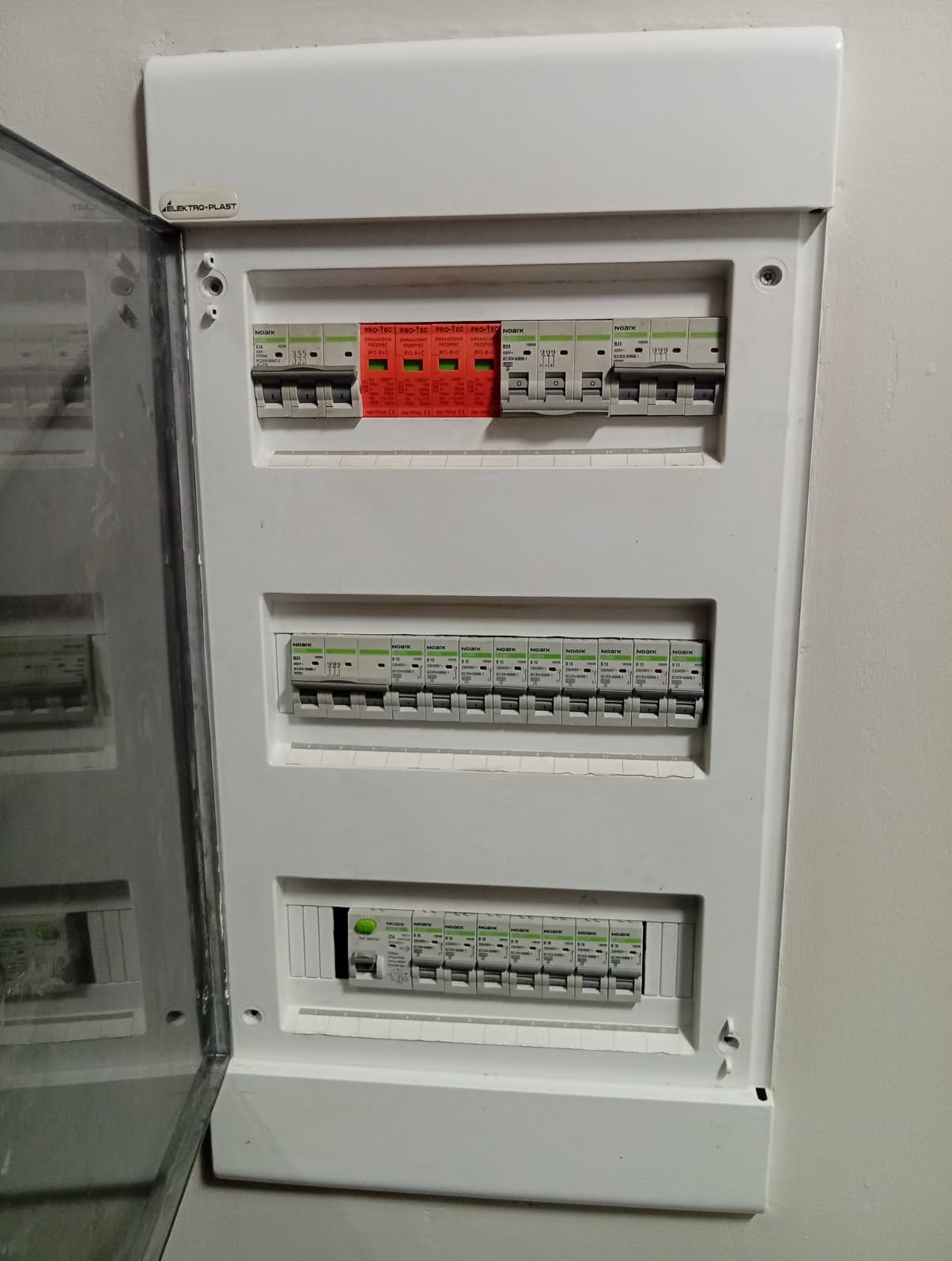Otwarta skrzynka elektryczna z bezpiecznikami i zabezpieczeniami przepięciowymi Noark i Elektro-Plast, zamontowana na ścianie. Widoczne trzy rzędy wyłączników.