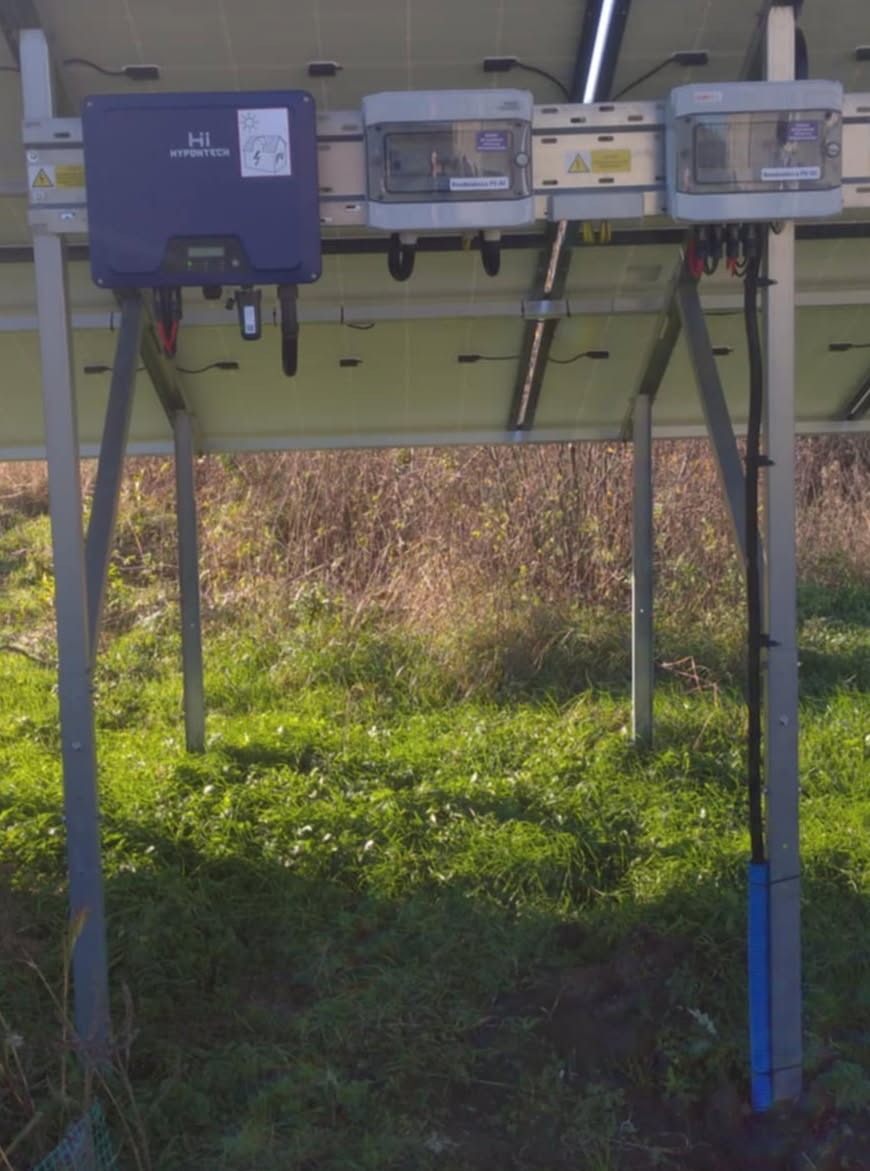 Instalacja fotowoltaiczna od spodu: widoczne panele, inwerter Hypontech i okablowanie na tle trawy i krzewów. Ujęcie z dołu podkreśla konstrukcję wsporczą.