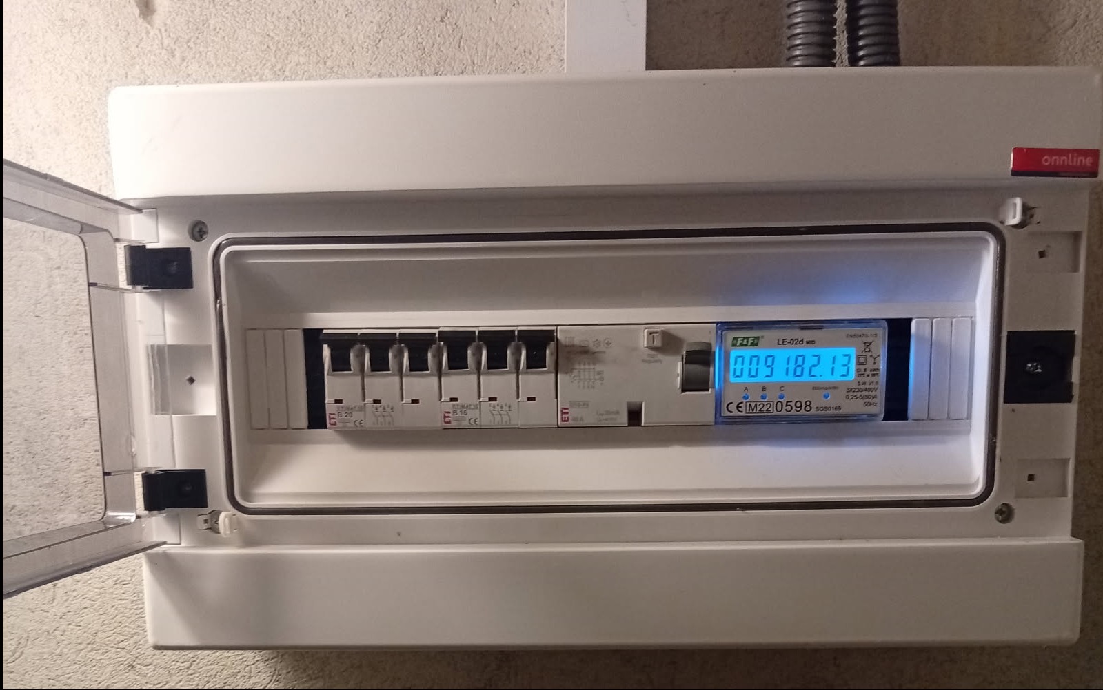 Otwarta skrzynka elektryczna z bezpiecznikami ETI i licznikiem F&F LE-02d MID, zamontowana na ścianie. Widoczne przewody w peszlu.