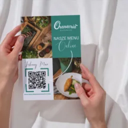 Dłonie trzymają ulotkę restauracji 'Oranżeria' z kodem QR i zdjęciami dań na tle białej tkaniny. Ulotka prezentuje menu online i zachęca do skanowania.