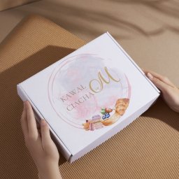 Klaudia Kozłowska Design - Białe pudełko z logo „Kawał Ciacha” w rękach na tle brązowej tektury falistej. Grafika z akwarelowymi ciastkami i różową obwódką. Delikatne światło.