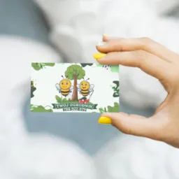 Dłoń z żółtym manicure trzyma wizytówkę z ilustracją dwóch pszczół, drzewa i kosiarki. Napis: Twój Ogrodnik, numer telefonu. Pastelowe tło.