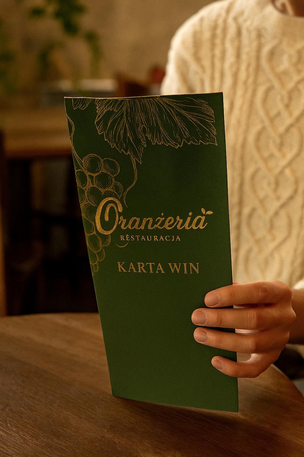 Karta win restauracji 'Oranżeria' w odcieniach zieleni i złota, trzymana w dłoniach nad drewnianym stołem. Elegancki design z motywem winogron.
