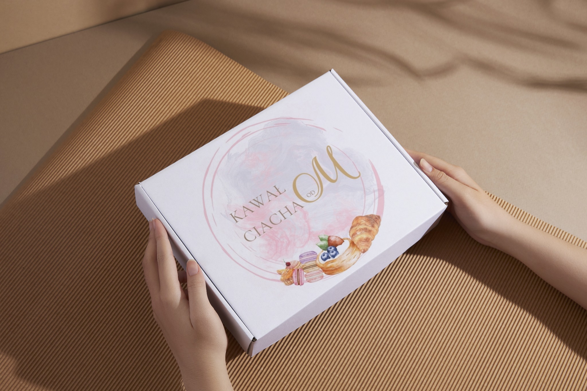 Białe pudełko z logo „Kawał Ciacha” w rękach na tle brązowej tektury falistej. Grafika z akwarelowymi ciastkami i różową obwódką. Delikatne światło.