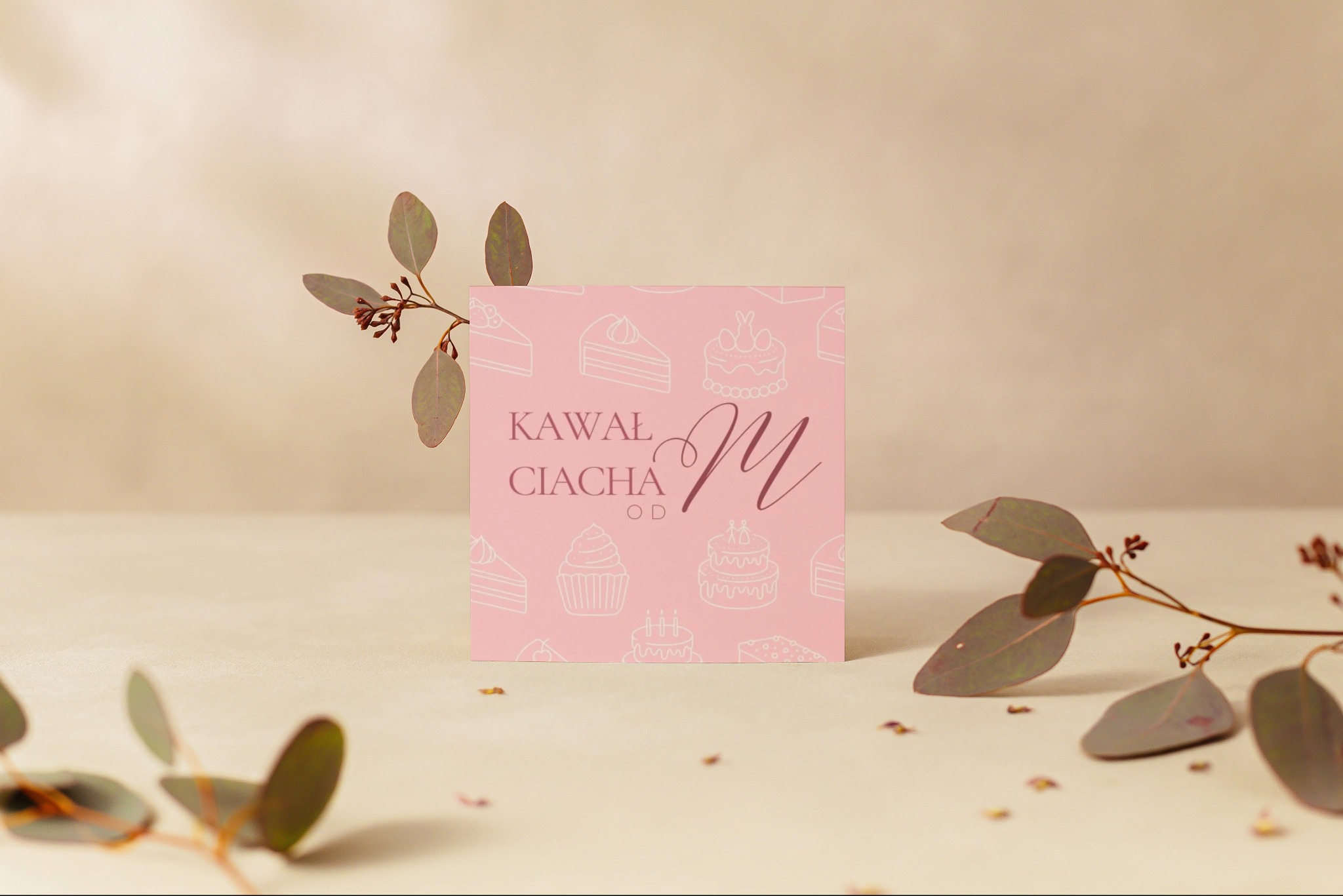 Różowa kartka z napisem 'Kawał Ciacha' i grafiką ciast, otoczona gałązkami eukaliptusa na beżowym tle.
