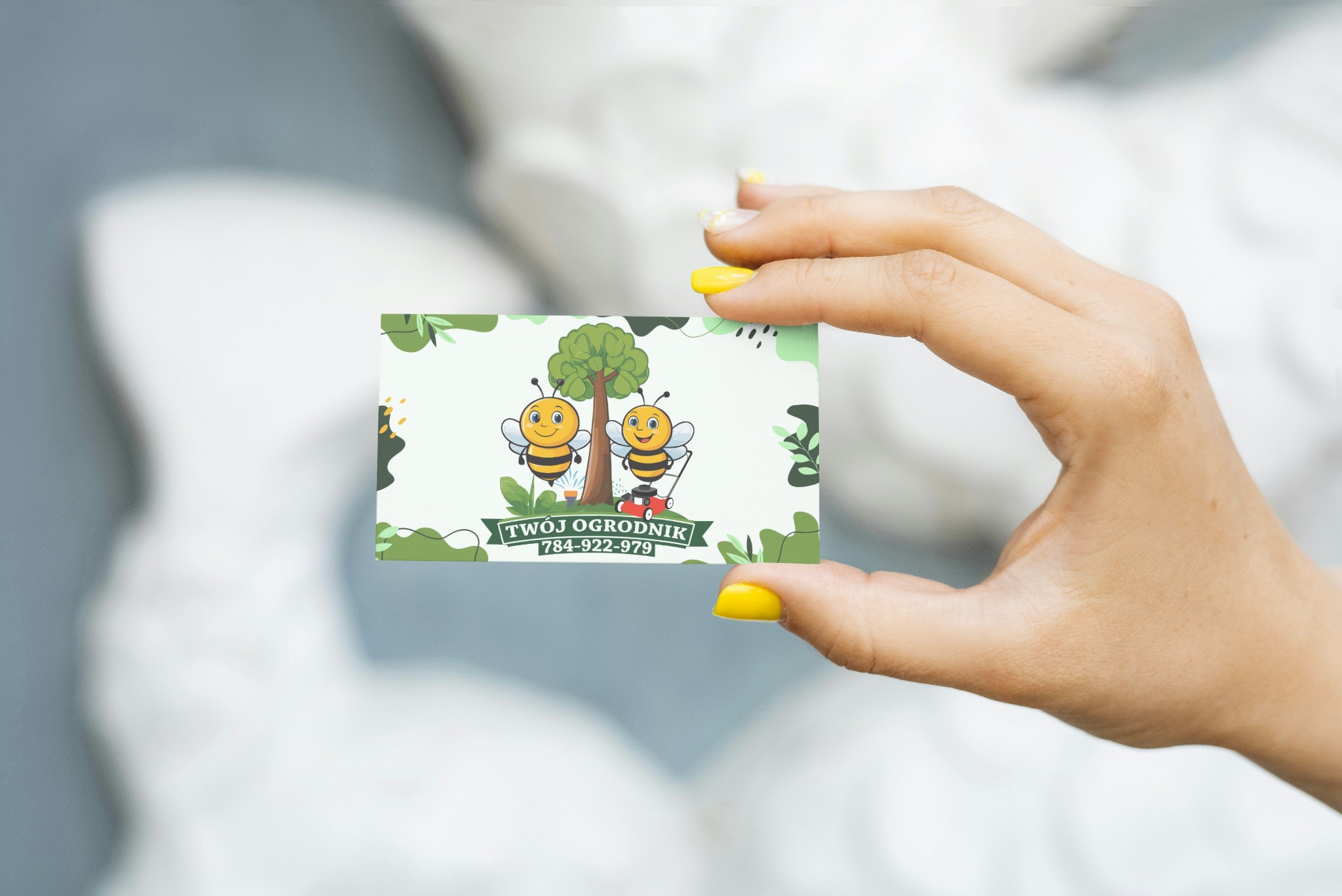 Dłoń z żółtym manicure trzyma wizytówkę z ilustracją dwóch pszczół, drzewa i kosiarki. Napis: Twój Ogrodnik, numer telefonu. Pastelowe tło.