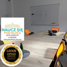 Sala lekcyjna z długim stołem, krzesłami z pomarańczowymi oparciami, białą tablicą z rysunkami i logo szkoły językowej 'Naucz Się Pod Klucz'.