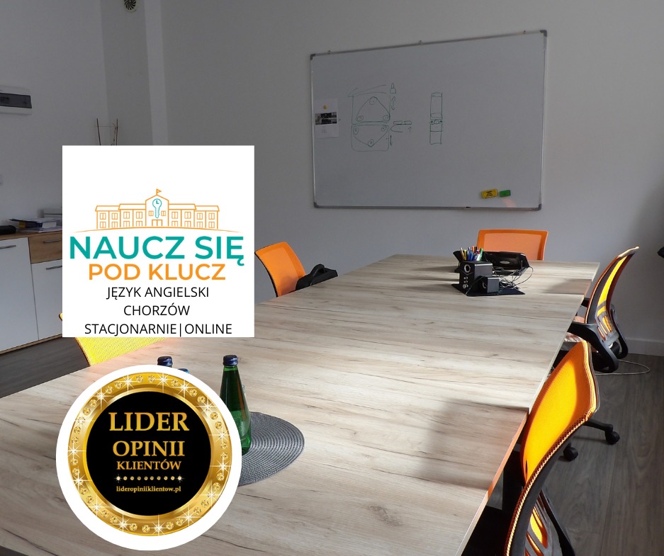 Sala lekcyjna z długim stołem, krzesłami z pomarańczowymi oparciami, białą tablicą z rysunkami i logo szkoły językowej 'Naucz Się Pod Klucz'.