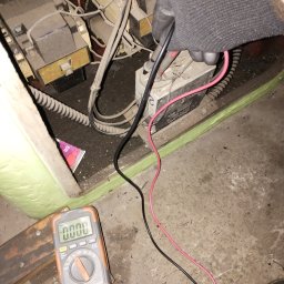 PRĄDPOMISTRZOWSKU - Elektryk w rękawicach ochronnych mierzy napięcie w skrzynce elektrycznej za pomocą multimetru. Widoczne przewody, złącza i elementy instalacji.