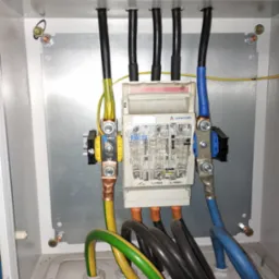 Rozdzielnica elektryczna z bezpiecznikami RBK 60 firmy Apator, widoczne kable w kolorach: czarnym, żółto-zielonym i niebieskim. Całość umieszczona w białej skrzynce.