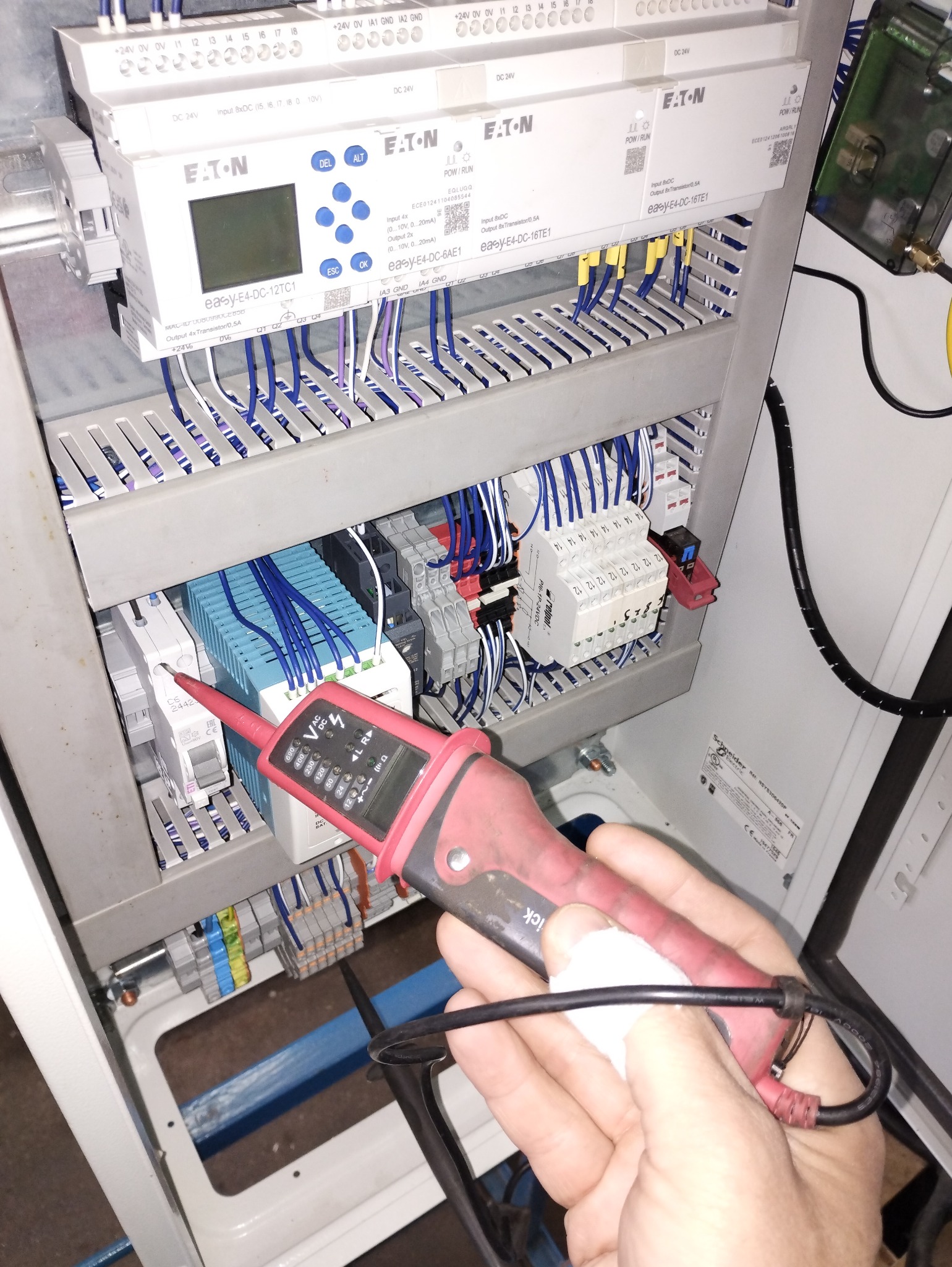 Pomiar napięcia w szafie sterowniczej. Tester napięcia w dłoni elektryka przy listwie zaciskowej z przewodami. Kontroler Eaton easy-E4-DC-12TC1 i inne moduły.