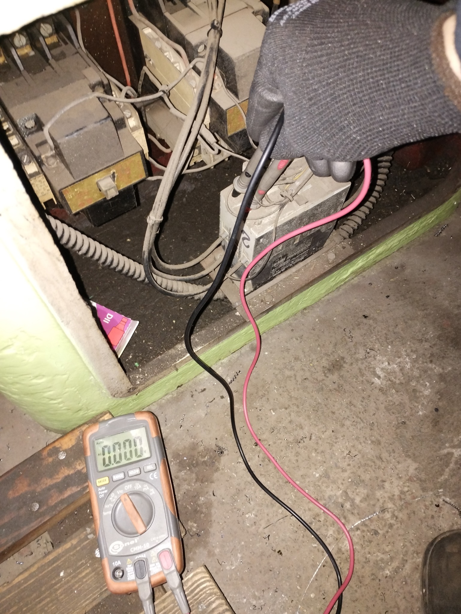 Elektryk w rękawicach ochronnych mierzy napięcie w skrzynce elektrycznej za pomocą multimetru. Widoczne przewody, złącza i elementy instalacji.