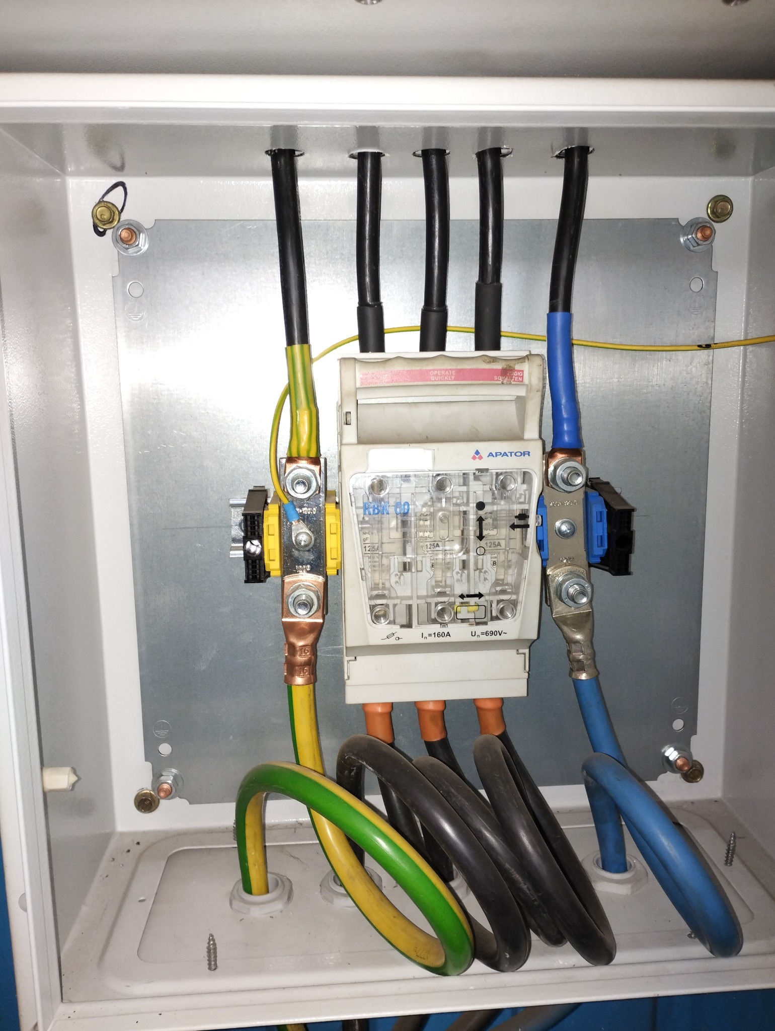 Rozdzielnica elektryczna z bezpiecznikami RBK 60 firmy Apator, widoczne kable w kolorach: czarnym, żółto-zielonym i niebieskim. Całość umieszczona w białej skrzynce.