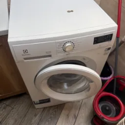 Biała pralka Electrolux z otwartymi drzwiczkami, widoczna w łazience z mopem. Ustawienia na panelu sterowania wskazują różne programy prania.