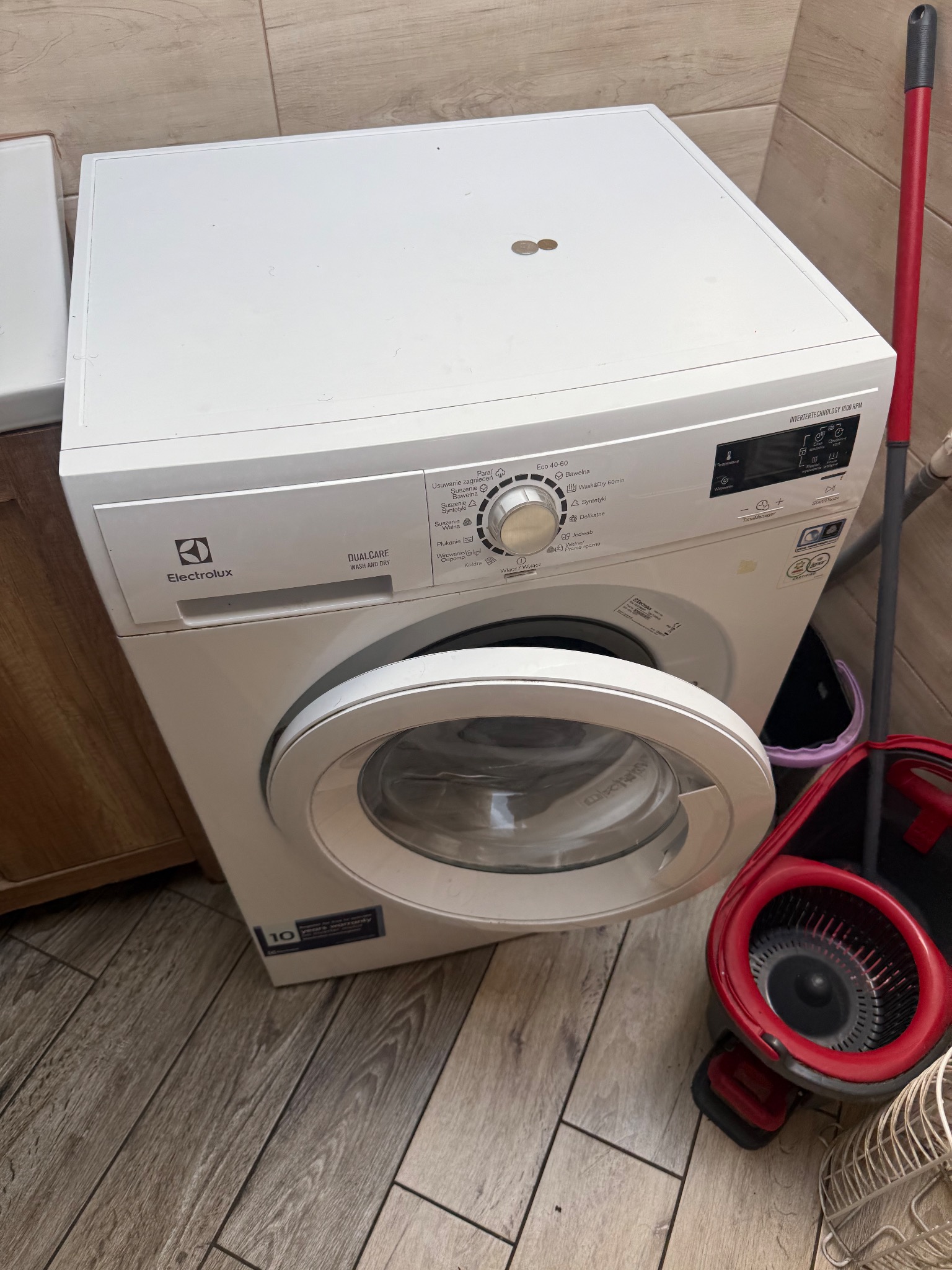 Biała pralka Electrolux z otwartymi drzwiczkami, widoczna w łazience z mopem. Ustawienia na panelu sterowania wskazują różne programy prania.