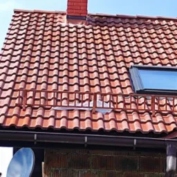 Czerwony dach z dachówki karpiówki z kominem z cegły klinkierowej i oknem dachowym, widok z dołu na tle błękitnego nieba. Widoczna antena satelitarna.