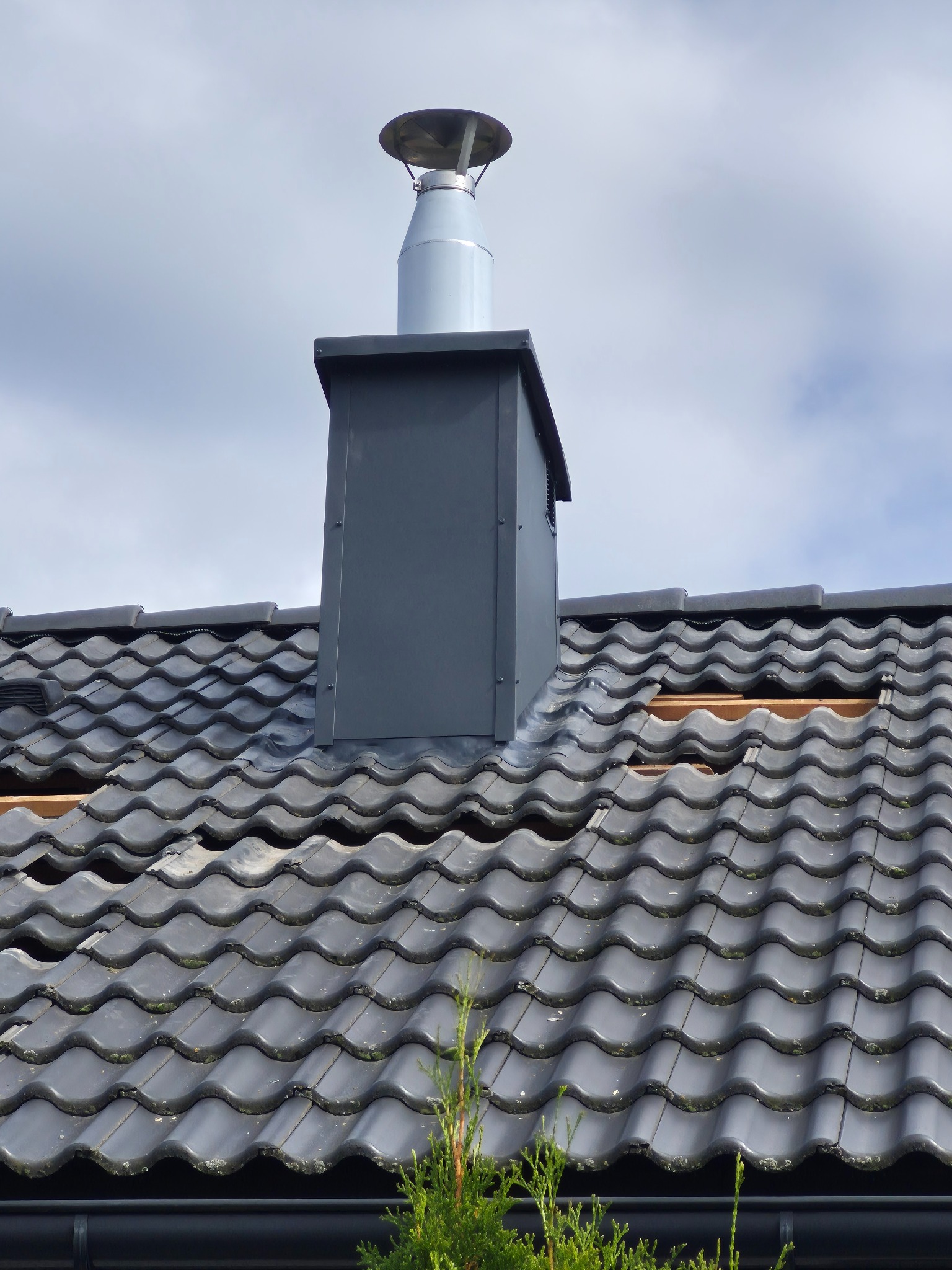 Szary komin wentylacyjny na dachu z ciemnej dachówki ceramicznej. Fragment dachu z widoczną łatą. Ujęcie z dołu, pochmurne niebo w tle.