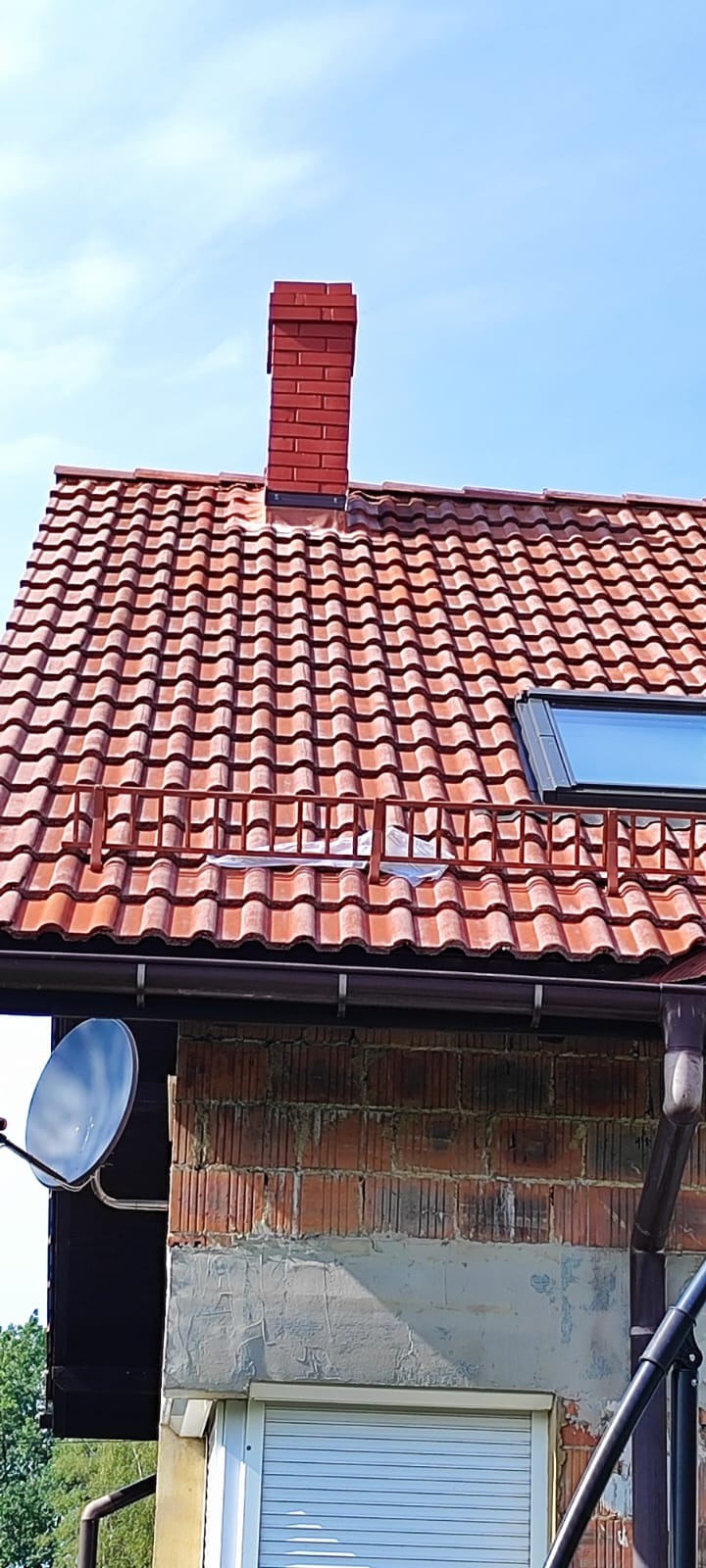 Czerwony dach z dachówki karpiówki z kominem z cegły klinkierowej i oknem dachowym, widok z dołu na tle błękitnego nieba. Widoczna antena satelitarna.