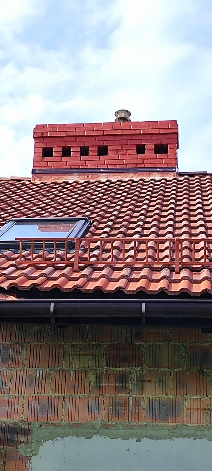 Ceglany komin z daszkiem wentylacyjnym nad dachem z dachówki ceramicznej z oknem połaciowym i barierką. Widoczna rynna i fragment ściany z cegły.