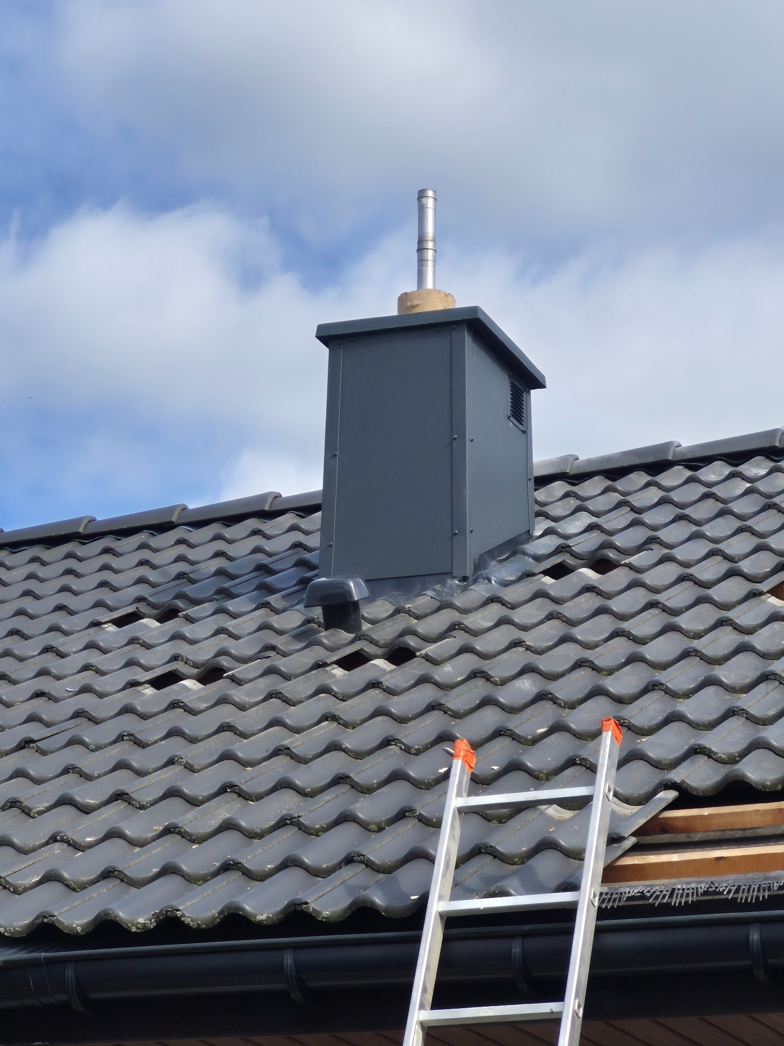Szary komin wentylacyjny na dachu z ciemnej dachówki ceramicznej. Fragment dachu uszkodzony, widoczna drabina. Błękitne niebo z chmurami w tle.