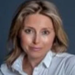 Portret kobiety w białej koszuli na ciemnym tle. Uśmiechnięta blondynka z delikatnym makijażem, patrzy wprost. Profesjonalny wizerunek.