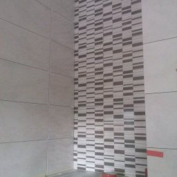 Adam Wierzbicki - Ściana w trakcie układania płytek z dekoracyjnym pasem mozaiki. Widoczne narzędzia glazurnicze i taśma zabezpieczająca. Prace wykończeniowe w łazience.