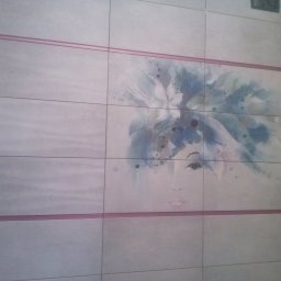 Adam Wierzbicki - Ściana wyłożona jasnymi płytkami z dekoracyjnym motywem kobiety w niebieskich odcieniach, oddzielona różową listwą. Widoczny narożnik pomieszczenia.