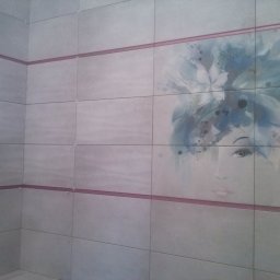 Adam Wierzbicki - Ściana wyłożona jasnymi płytkami z dekoracyjnym motywem kobiety w stylu akwareli, oddzielona bordową listwą. Widoczny narożnik pomieszczenia.