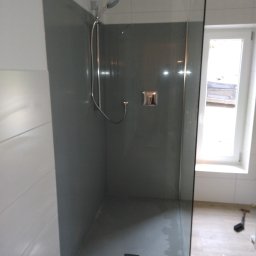 Adam Wierzbicki - Nowoczesna kabina prysznicowa walk-in z szarą ścianą i szklanym panelem. Widoczny odpływ liniowy i okno w tle. Minimalistyczny design.