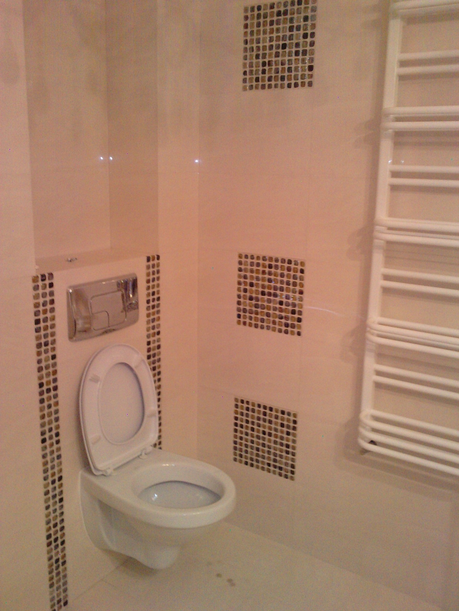 Wykończona łazienka z podwieszaną toaletą, obudowaną mozaiką. Na ścianie dekoracyjne kwadraty z mozaiki i biały grzejnik drabinkowy. Jasne płytki na ścianach.