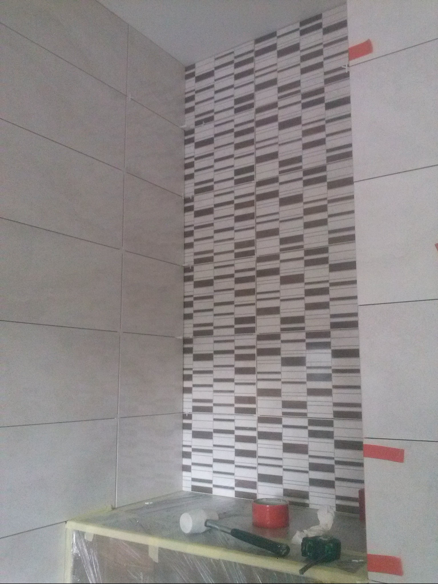 Ściana w trakcie układania płytek z dekoracyjnym pasem mozaiki. Widoczne narzędzia glazurnicze i taśma zabezpieczająca. Prace wykończeniowe w łazience.