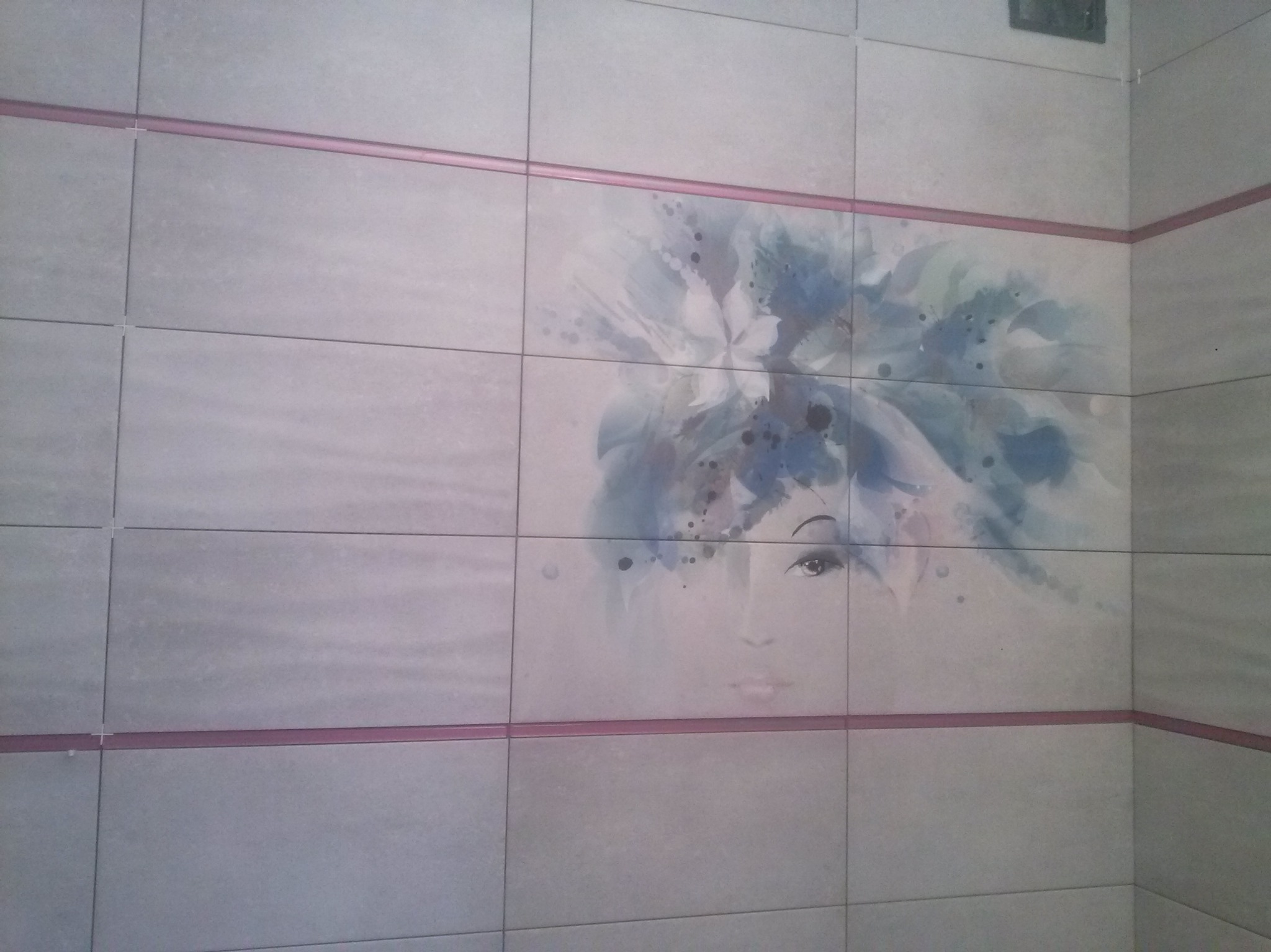 Ściana wyłożona jasnymi płytkami z dekoracyjnym motywem kobiety w niebieskich odcieniach, oddzielona różową listwą. Widoczny narożnik pomieszczenia.