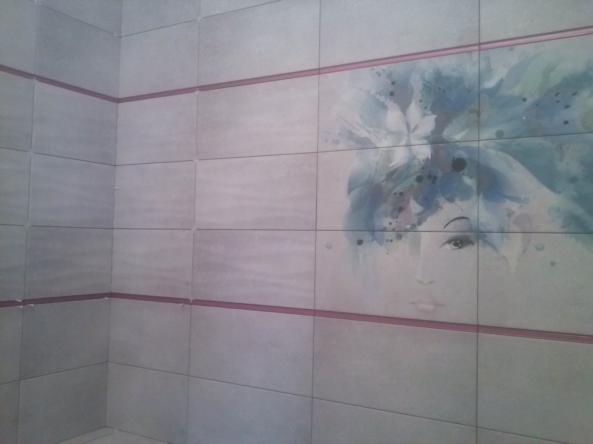 Ściana wyłożona jasnymi płytkami z dekoracyjnym motywem kobiety w stylu akwareli, oddzielona bordową listwą. Widoczny narożnik pomieszczenia.