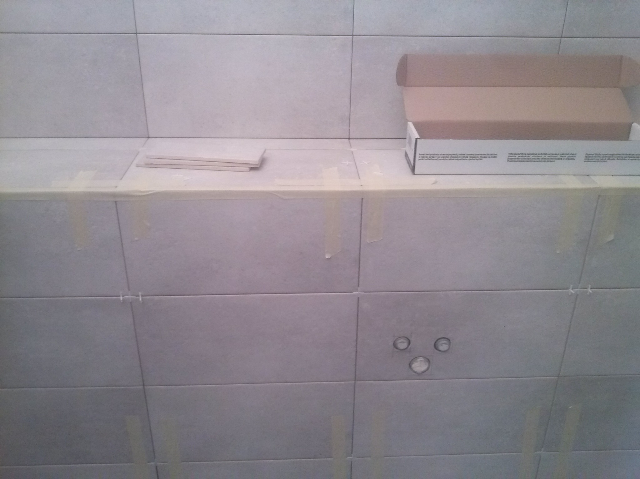 Ściana w łazience z szarymi płytkami, zabezpieczona taśmą malarską. Widoczne krzyżyki dystansowe, otwory na instalacje i karton po płytkach.