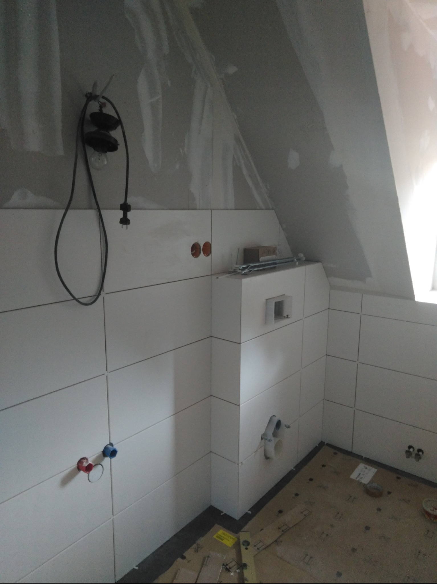 Białe płytki w łazience na poddaszu, widoczne otwory na instalacje, przygotowane do montażu armatury. Częściowo wykończone ściany, surowy stan podłogi.