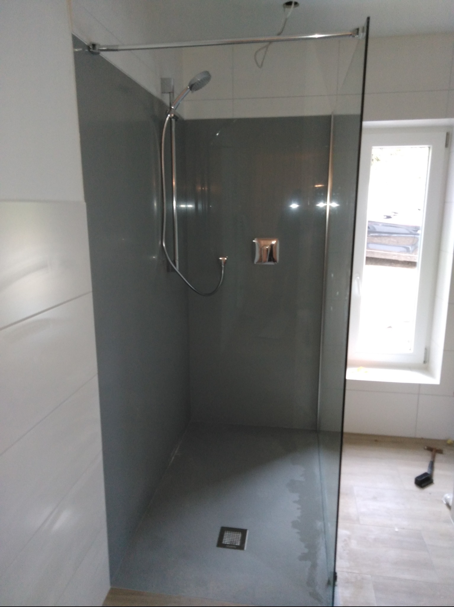 Nowoczesna kabina prysznicowa walk-in z szarą ścianą i szklanym panelem. Widoczny odpływ liniowy i okno w tle. Minimalistyczny design.