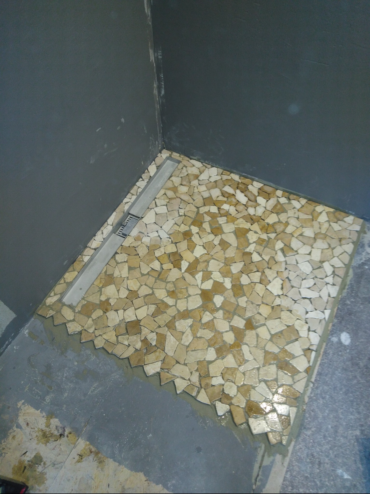 Mozaika z kamieni w odcieniach beżu i brązu ułożona na podłodze prysznica z odpływem liniowym. Szare ściany w tle. Widok z góry.