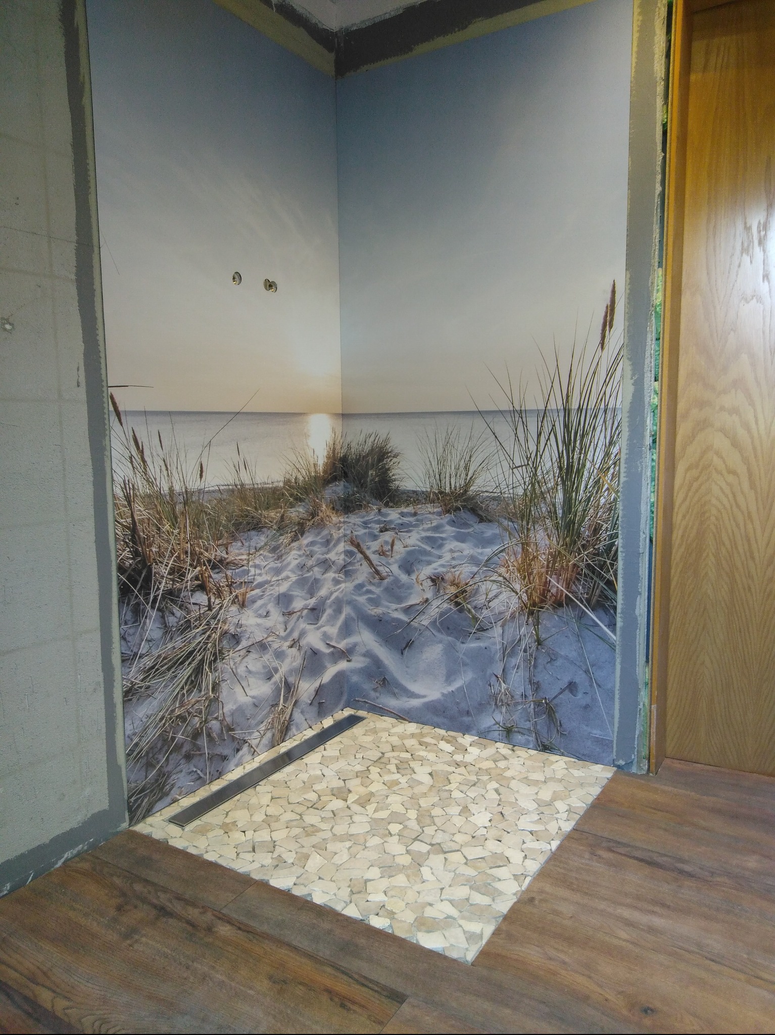 Wykończona strefa prysznicowa z odpływem liniowym i mozaiką z kamyków, ściana z fototapetą plaży. Podłoga z paneli drewnopodobnych.