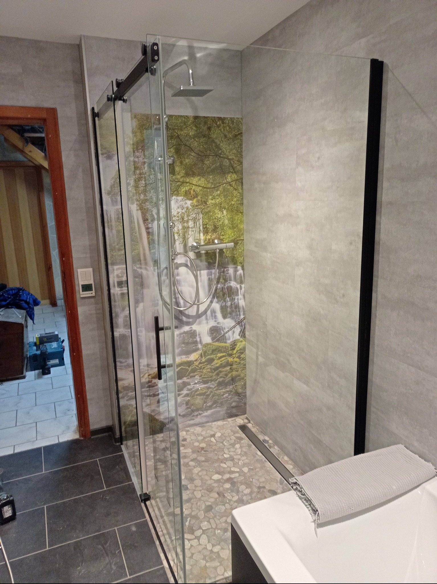 Nowoczesna kabina prysznicowa z przesuwnymi drzwiami, wykończona szarą płytką imitującą kamień i panelem z motywem wodospadu. Widoczny odpływ liniowy i fragment wanny.