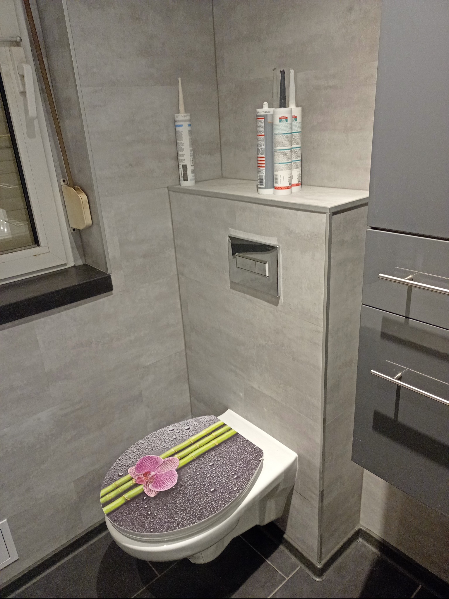 Szara łazienka po remoncie: płytki imitujące kamień, podwieszana toaleta z ozdobną deską, szara szafka i silikony na zabudowie. Nowoczesne wykończenie.