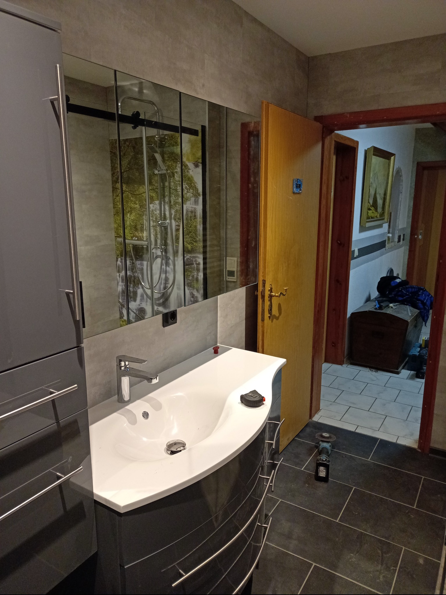 Nowoczesna łazienka z szarą szafką pod umywalką i lustrem z motywem wodospadu. Na podłodze ciemne płytki. Widoczna szlifierka kątowa.