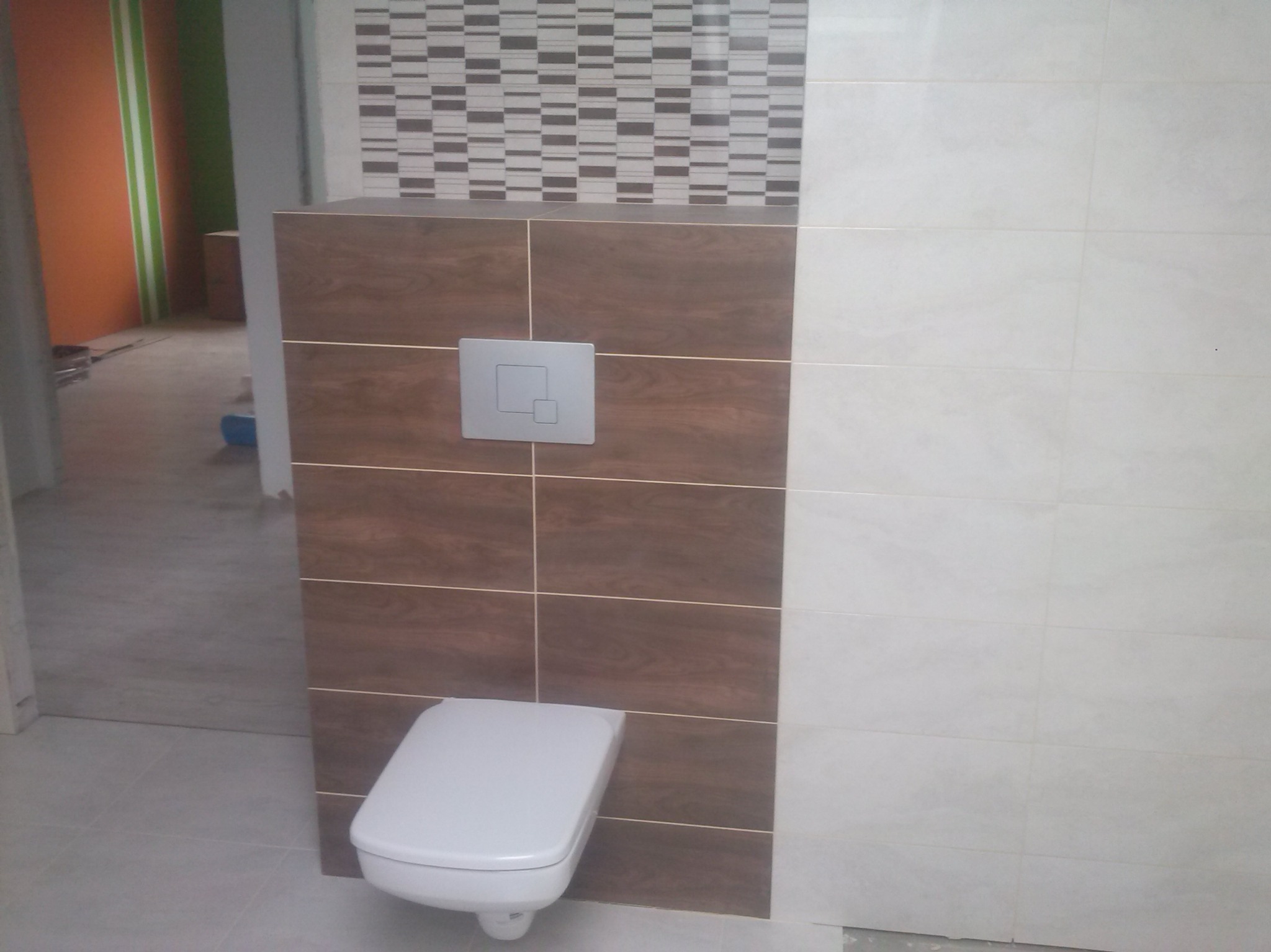 Podwieszana toaleta z brązowymi i białymi płytkami w łazience. Widoczne mozaiki na górze. Świeżo wyremontowane wnętrze z pomarańczowymi ścianami w tle.