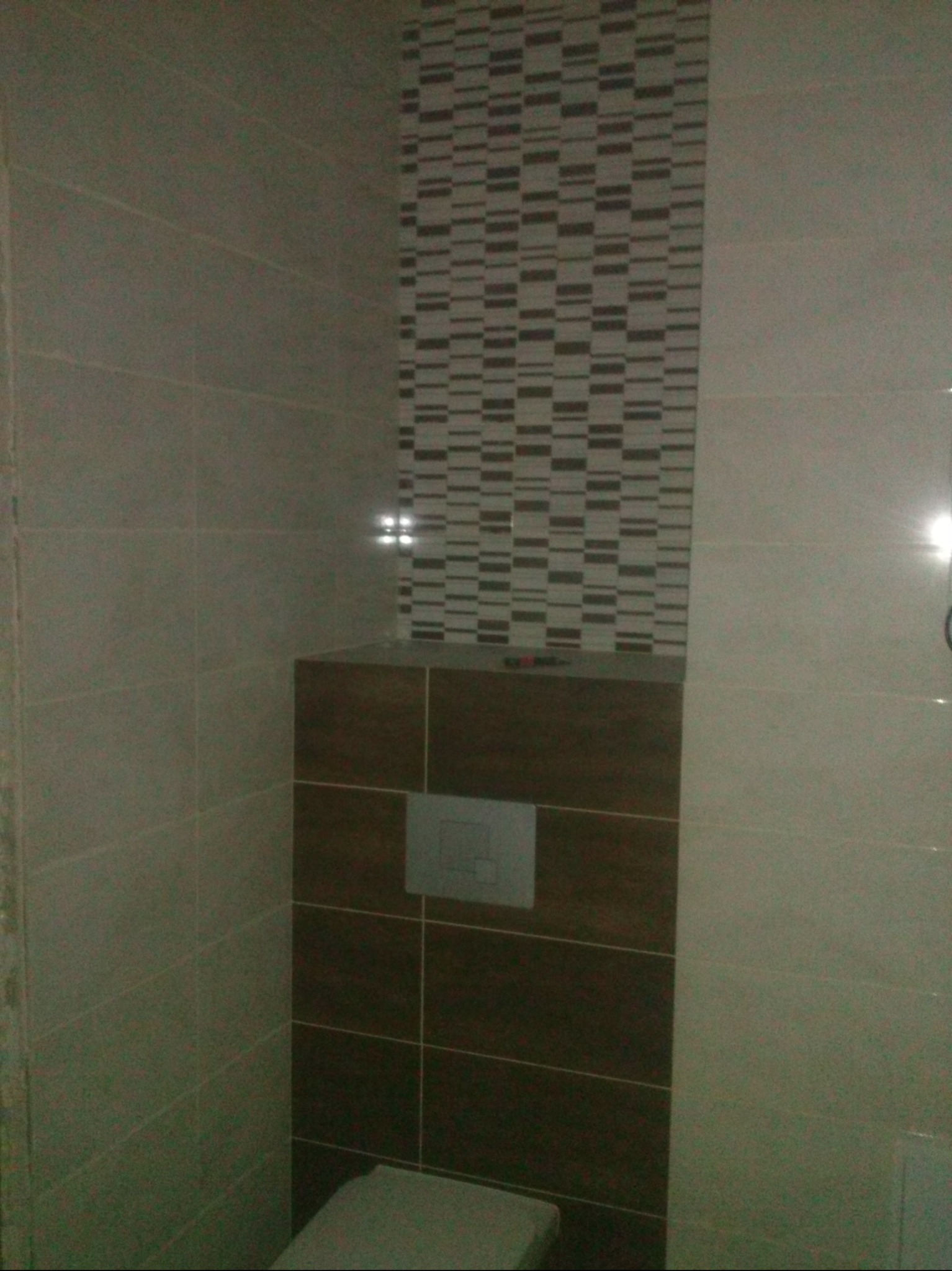 Wykończona toaleta z brązowymi i beżowymi płytkami, mozaika na ścianie. Widoczny przycisk spłuczki i fragment muszli. Minimalistyczny design.