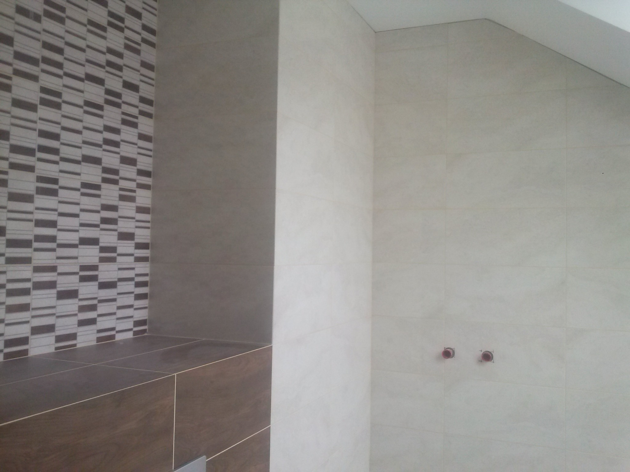 Wykończona ściana łazienki z beżowych płytek, fragment dekoracyjnej mozaiki i zabudowa z ciemnobrązowych płytek. Widoczne przyłącza wodne.