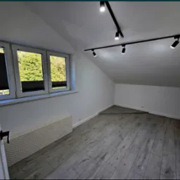 Pok&oacute;j na poddaszu po remoncie: szare panele, białe ściany, okno z żaluzjami i nowoczesne oświetlenie sufitowe. Minimalistyczny design.